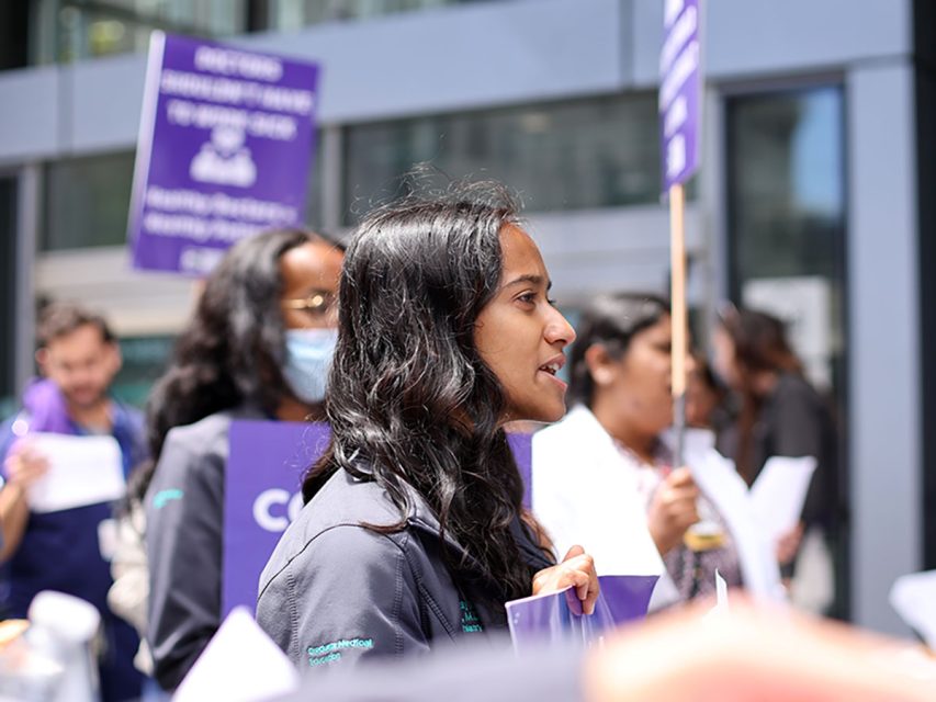A group of healthcare workers in professional attire hold signs and leaflets during a daylight protest outside a modern building. One central figure appears to be speaking or addressing the group.