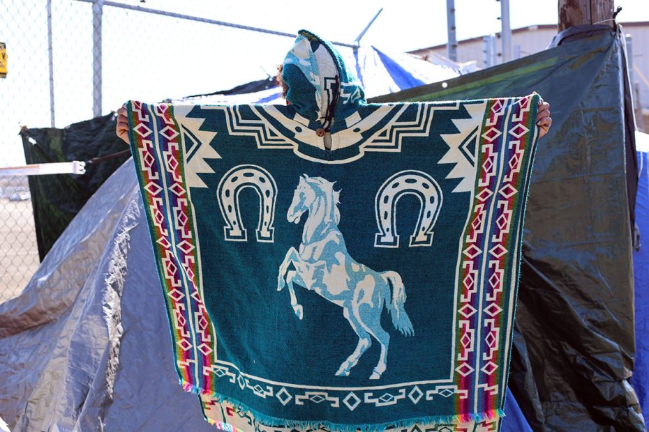 Person holding up a poncho with a rearing horse and horseshoe design, standing in front of a chain-link fence and tarpaulin.