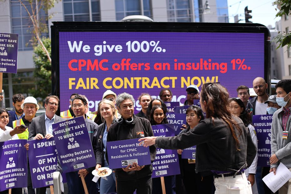 A group of people stand in front of a large sign that reads, "We give 100%. CPMC offers an insulting 1%! FAIR CONTRACT NOW!" Some hold signs advocating for fair contracts.