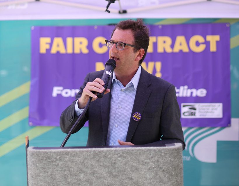A man in a suit speaks into a microphone at a podium. A sign behind him reads "FAIR CONTRACT NOW!.