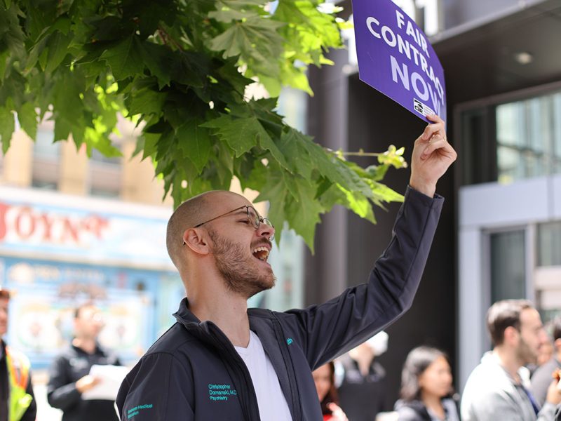 A man wearing glasses and a jacket holds a "Fair Contract Now" sign during an outdoor protest. Others are gathered in the background, and large green leaves from a tree are visible above him.