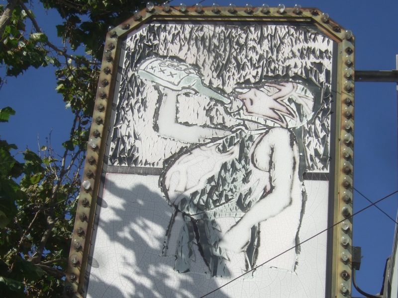 A mural depicts an abstract figure in a tank top drinking from a bottle. The artwork is mounted on an outdoor structure, framed with bulbs, and bordered by foliage and a blue sky.