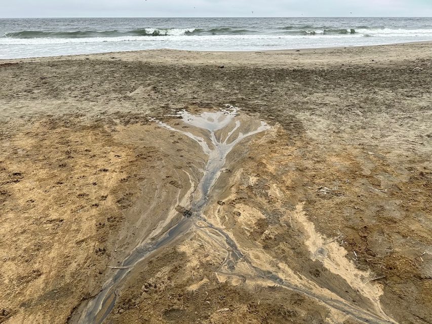 A small stream of water flows through a sandy beach towards the ocean, with gentle waves in the background under a cloudy sky.