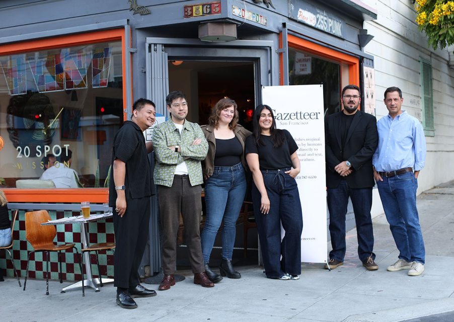 A group of six people are standing and smiling in front of an orange-framed storefront with a "20 Spot" sign. A tabletop, chairs, and a stand-up sign for "Gazetteer" are also visible.