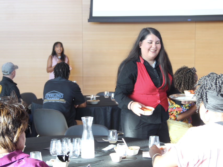 A server in a red vest holds a tray while smiling at a seated guest. In the background, a person speaks at a podium in front of a screen. Other people are seated at tables at the Taste of Bayview event.