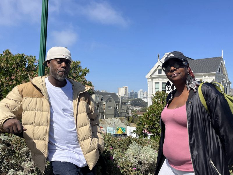 Two individuals stand outside on a sunny day with buildings and greenery in the background. One person wears a beige jacket and white cap, the other wears a pink top, black jacket, and black cap.