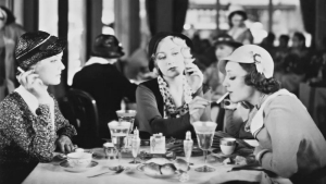 Three women sit at a table in a restaurant, drinking and smoking, with drinks and food in front of them. Other patrons are seated in the background.