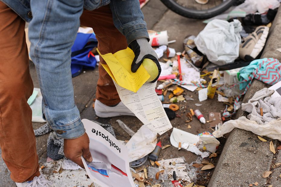 A person wearing gloves and a denim jacket sweeps the littered street, collecting papers amid scattered trash and debris.