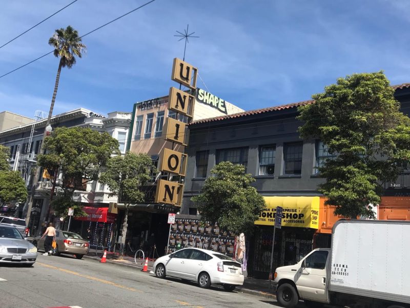 Street view of a building with a large vertical "UNION" sign in front. There are shops, cars, and pedestrians along the street, with trees lining the sidewalk.