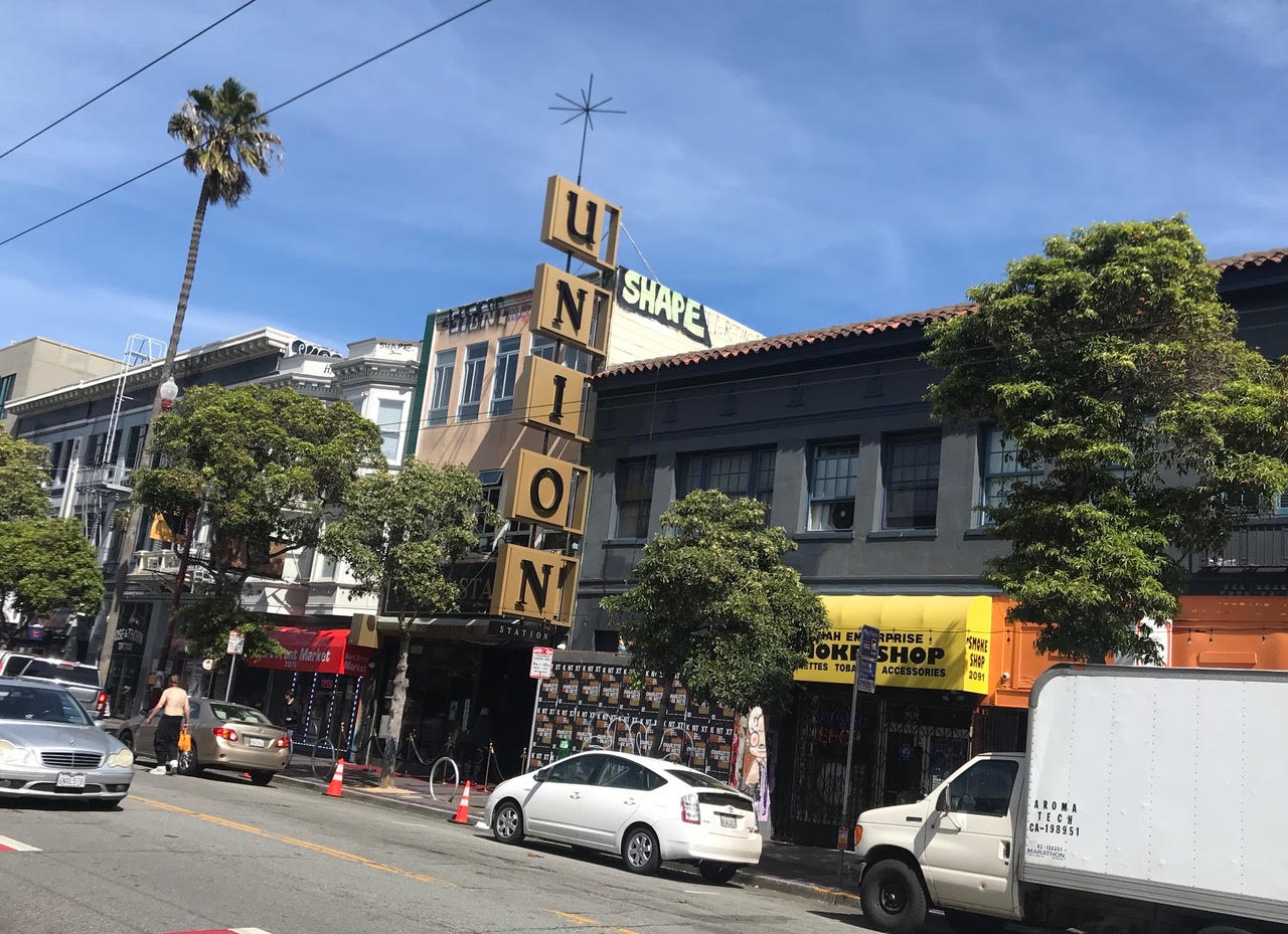 Street view of a building with a large vertical "UNION" sign in front. There are shops, cars, and pedestrians along the street, with trees lining the sidewalk.