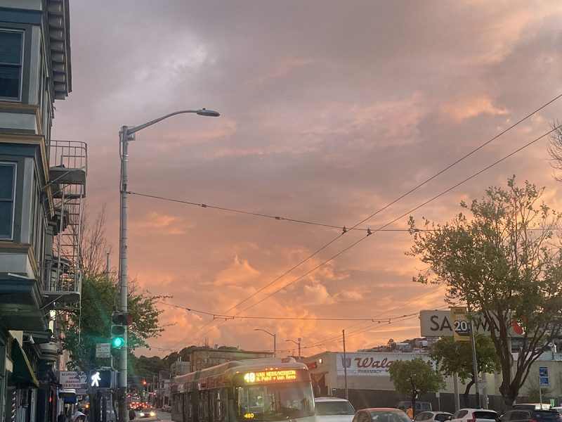 City street at sunset with cars, a bus, and pedestrians. The sky is filled with colorful clouds, and a green traffic light is visible. Buildings and trees line the street.