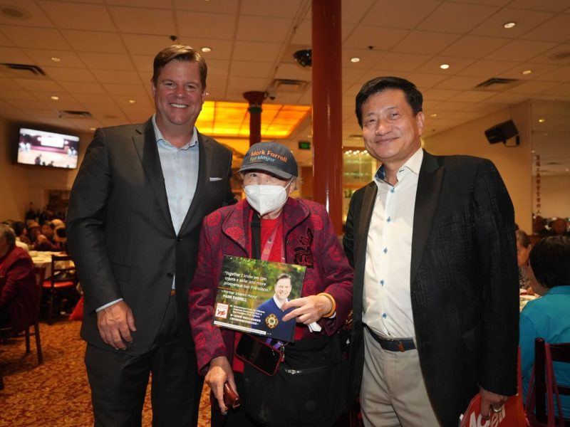 Three people stand inside a large room; two men, both in suits, flank an elderly woman wearing a mask and holding a sign. The room is crowded, with people seated at tables in the background.