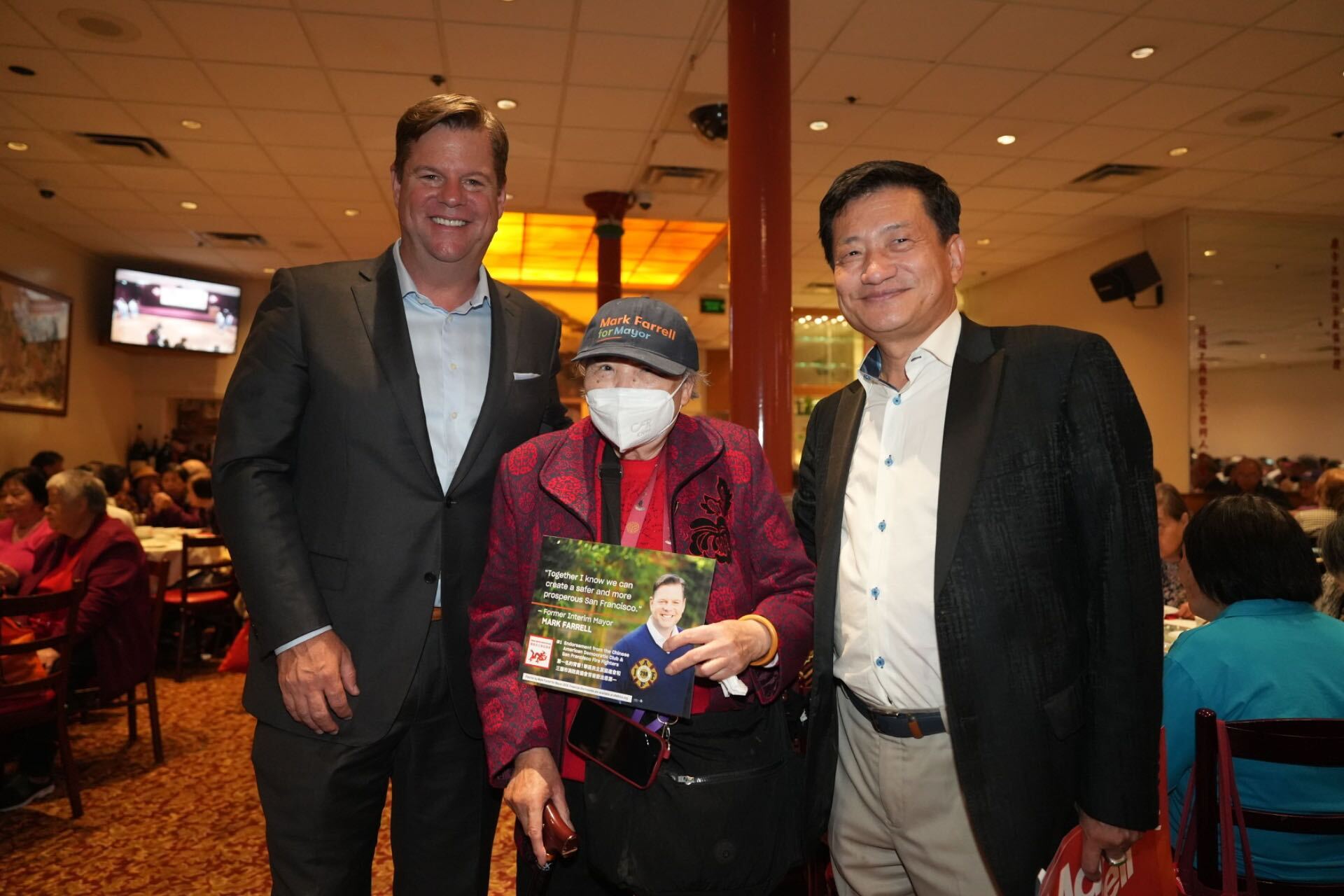 Three people stand inside a large room; two men, both in suits, flank an elderly woman wearing a mask and holding a sign. The room is crowded, with people seated at tables in the background.