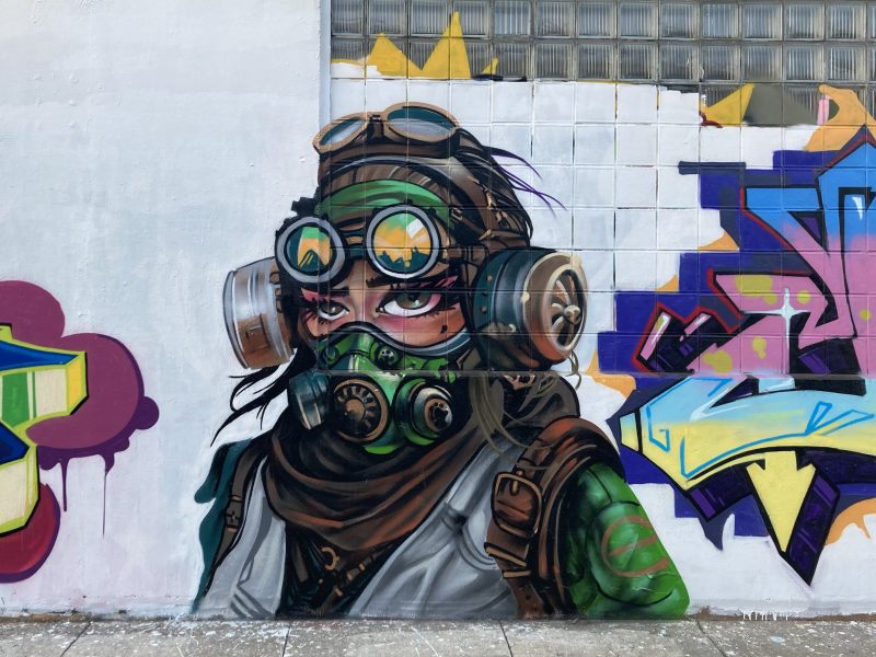 A mural of a person wearing goggles and a gas mask with a green and brown outfit, set against a wall with abstract mural art.