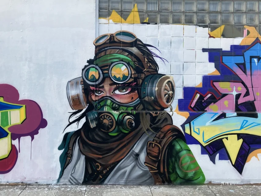 Wall painting of a person with green goggles, a respirator mask, headphones, and a scarf. The graffiti background includes geometric and abstract shapes in various colors like purple, blue, and yellow.