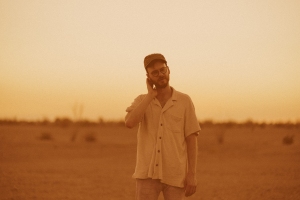A person wearing a cap and button-up shirt stands in an expansive desert landscape at sunset, with their hand on their neck.