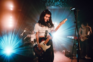 A guitarist smiles while playing an electric guitar on stage with bright lights shining in the background. A drummer and another guitarist are visible in the background.