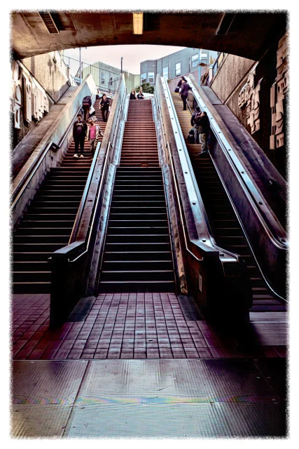 A staircase flanked by two escalators in a subway station. People are ascending and descending, and the background shows an open area with buildings and additional passengers.