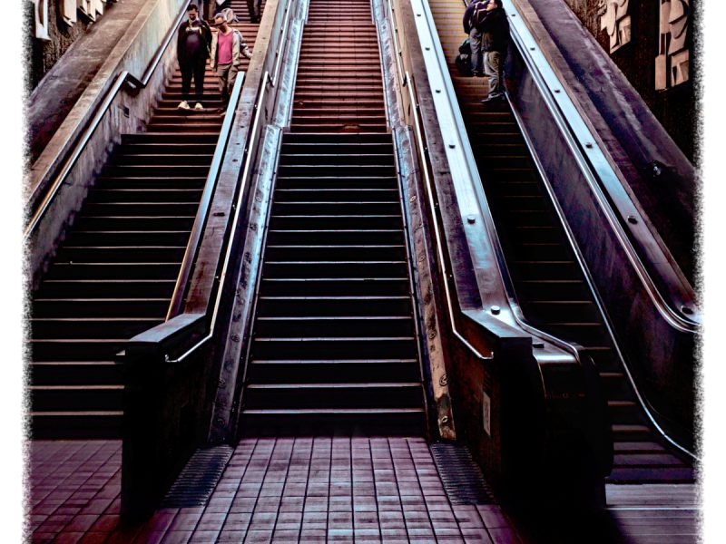 A flight of stairs and two escalators leading to an upper level, with several people ascending and descending, located in an outdoor urban setting.