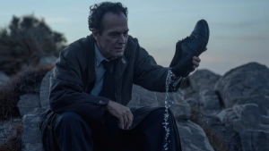 A man sitting on rocks in a suit pours water out of a shoe at dusk.