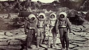 Four astronauts in space suits stand on a cracked, rocky terrain with mountains in the background and a large piece of equipment behind them. The image is in black and white.