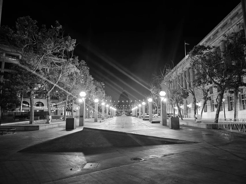 A black and white photo of an empty, well-lit urban plaza at night, lined with trees and flanked by buildings on both sides, with a dome visible in the background.