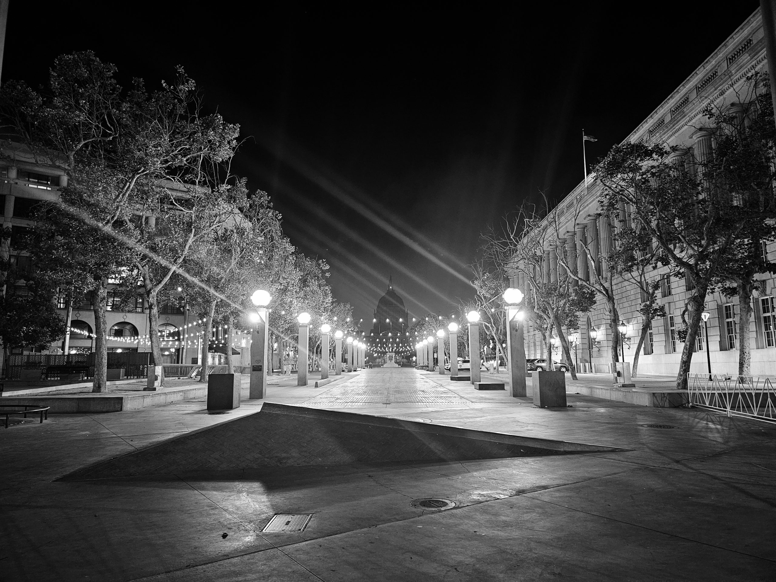 Black and white night scene of a deserted plaza flanked by trees and buildings with streetlights illuminating the pathway and a domed structure in the background.