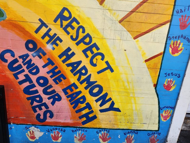 Mural with the text "RESPECT THE HARMONY OF THE EARTH AND OUR CULTURES" surrounded by handprints labeled with names against a colorful background.
