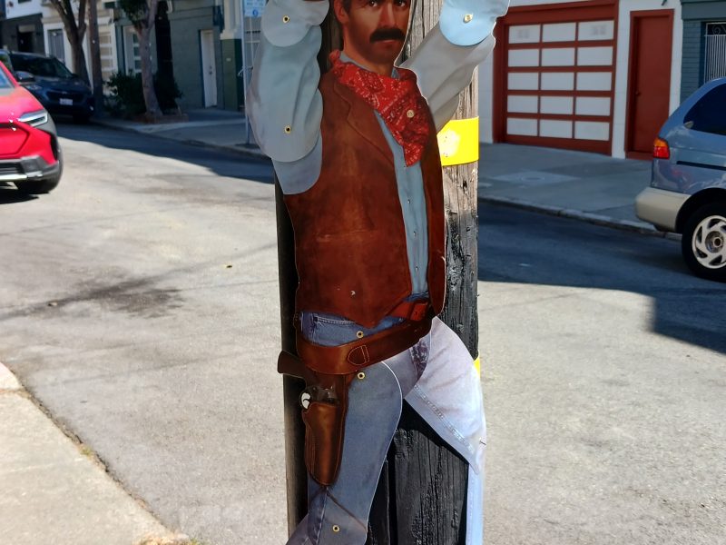 Wooden cutout of a cowboy with raised hands attached to a utility pole on a residential street with houses and vehicles in the background.