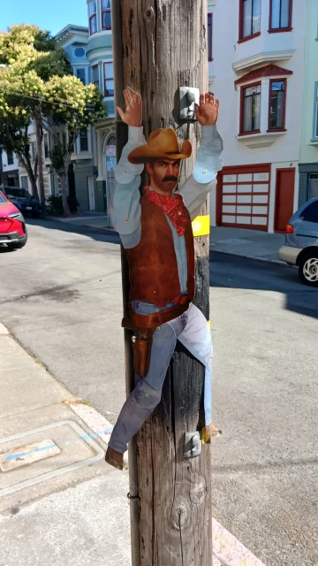 A cardboard cutout of a cowboy with raised arms is attached to a wooden utility pole on a residential street.