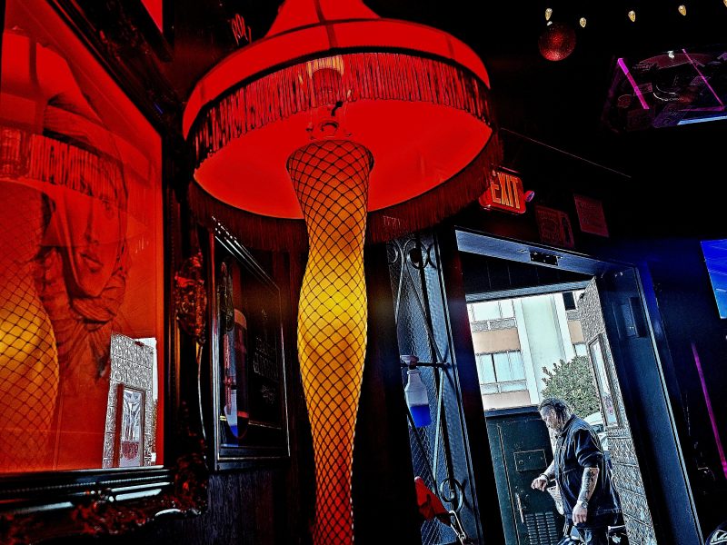 A leg-shaped lamp with a fringed shade is lit in a dimly lit bar, casting an orange glow. A person outside in the background is seen through the open doorway.