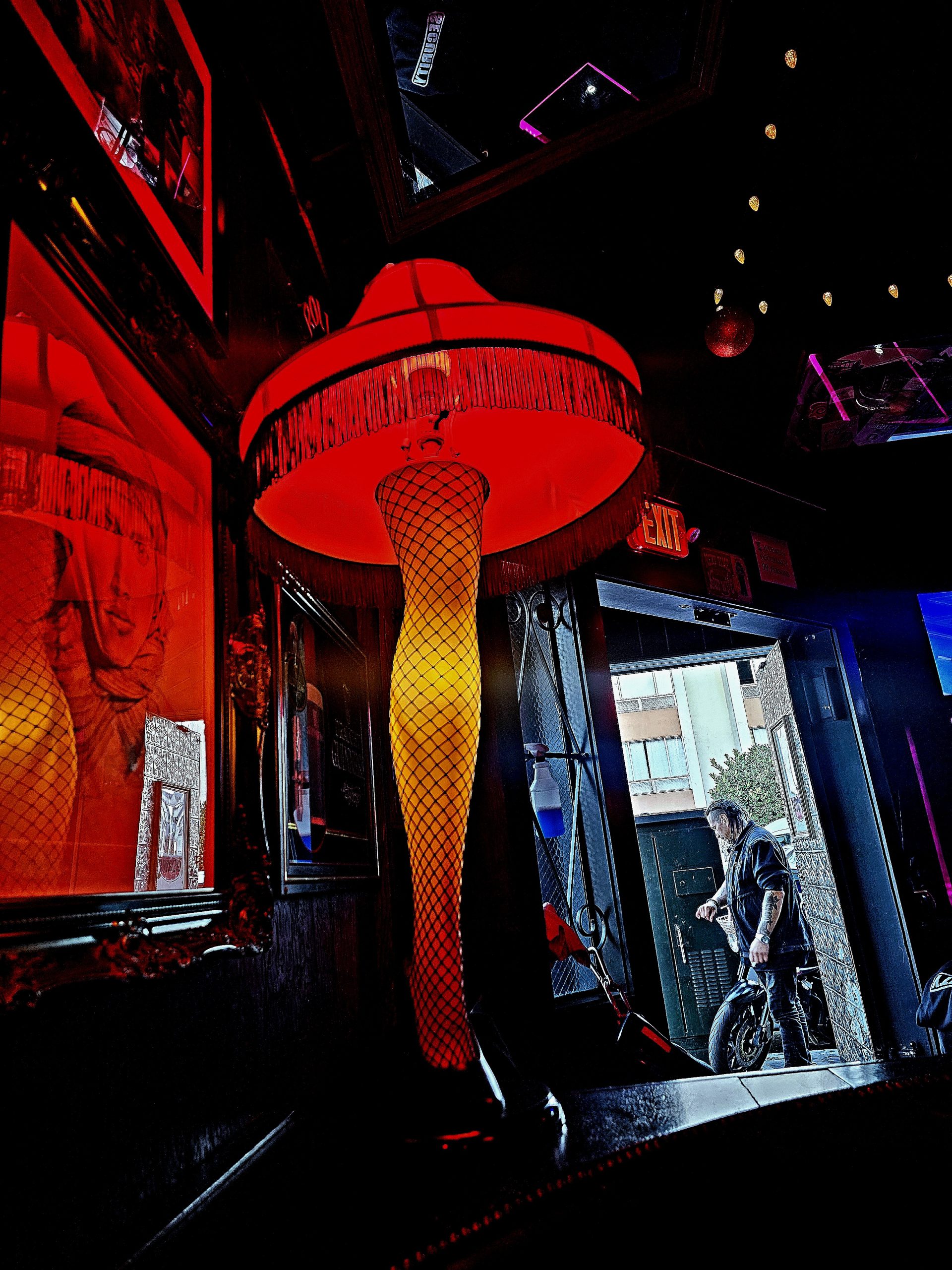 A leg-shaped lamp with a fringed shade is lit in a dimly lit bar, casting an orange glow. A person outside in the background is seen through the open doorway.