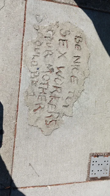 A section of a sidewalk with a patch of concrete containing the carved message: "Be nice to sex workers, your mother is one.