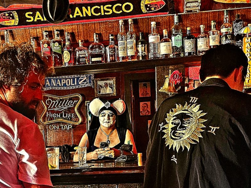 Three people in a bar. One woman is dressed in an elaborate costume behind the bar. Shelves of liquor bottles and a "San Francisco" sign are visible in the background.
