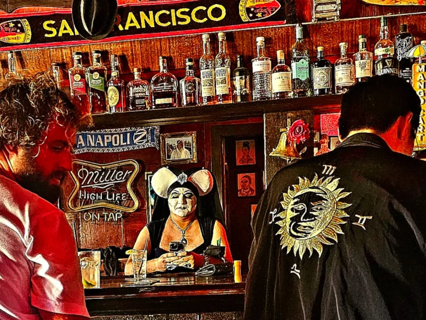 Three people in a bar with various liquor bottles on shelves; one person wearing a black outfit stands behind the bar, and two patrons are in the foreground. A "San Francisco" banner is displayed above.