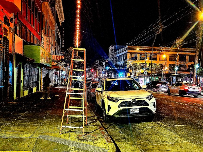 A white SUV is parked on a city street at night next to a tall ladder. Neon lights illuminate the scene. There are buildings and a few people in the background.