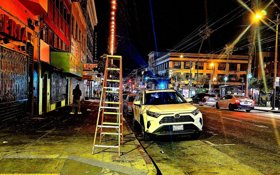 A white SUV is parked on a city street at night next to a tall ladder. Neon lights illuminate the scene. There are buildings and a few people in the background.