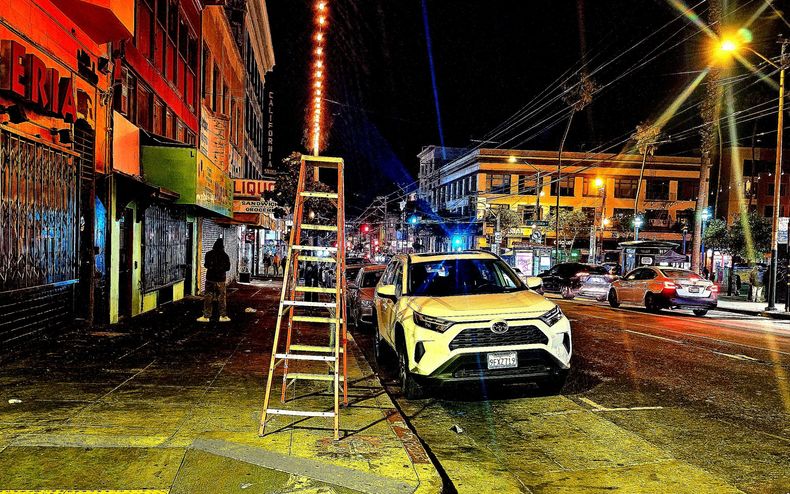 A white SUV is parked on a city street at night next to a tall ladder. Neon lights illuminate the scene. There are buildings and a few people in the background.