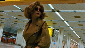A person wearing sunglasses and a trench coat walks through a spacious, well-lit indoor area with columns and ceiling lights.