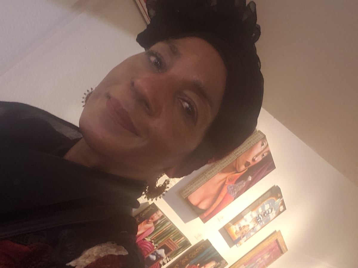 A person wearing a black headscarf and earrings smiles at the camera. Several colorful paintings are displayed on the wall in the background.