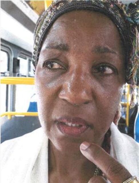 A close-up of a person looking to the side on a bus, with their finger pointing to their lips. They are wearing a headscarf.