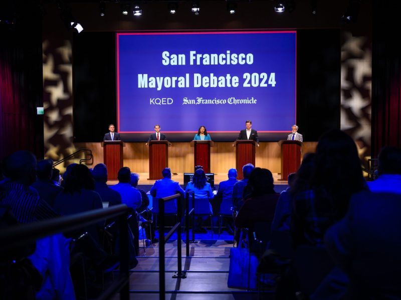 Five candidates stand behind podiums on stage during the San Francisco Mayoral Debate 2024, with an audience seated in front. The event is hosted by KQED and the San Francisco Chronicle.
