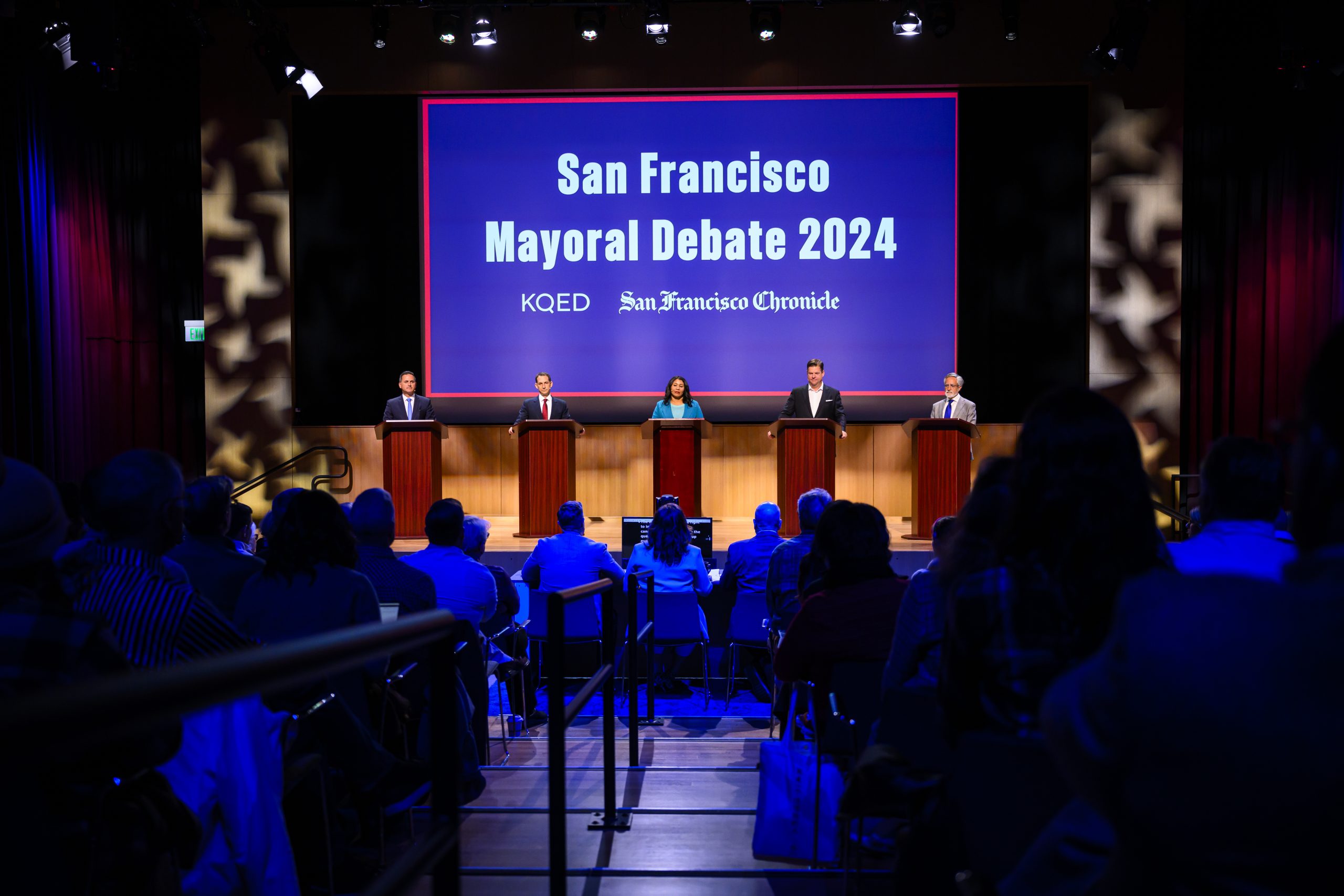 Five candidates stand behind podiums on stage during the San Francisco Mayoral Debate 2024, with an audience seated in front. The event is hosted by KQED and the San Francisco Chronicle.