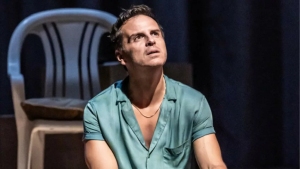 A person in a green shirt sits indoors, looking upward with a thoughtful expression. A white plastic chair is visible in the background.