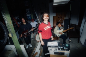 A four-person band performs in a cluttered room. The front person wears sunglasses and a red 'Cinci' shirt, with others playing guitar and bass in the background. Equipment and instruments are scattered around.
