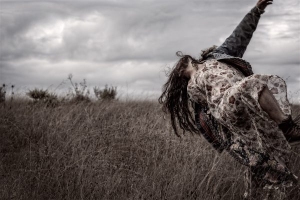 A figure in a floral dress leans back dramatically in a field under a cloudy sky, with an arm stretched upward.