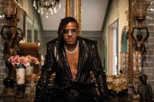 A person with sunglasses and a striped black outfit sits on a counter in a lavish room with ornate decor, including flowers, statues, and a large mirror.