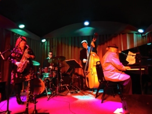 A jazz quartet performs on stage; a saxophonist, a drummer, an upright bassist, and a pianist play under colorful lighting.