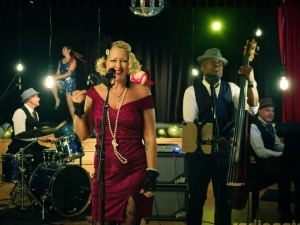 A five-member jazz band is performing on stage. The female singer is in front wearing a red dress, with the drummer, bassist, guitarist, and a dancer visible in the background.