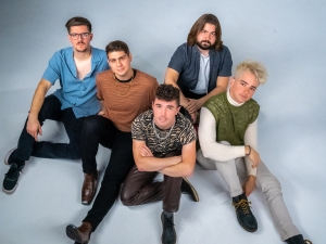 Five men are sitting on the floor against a light background, facing the camera.